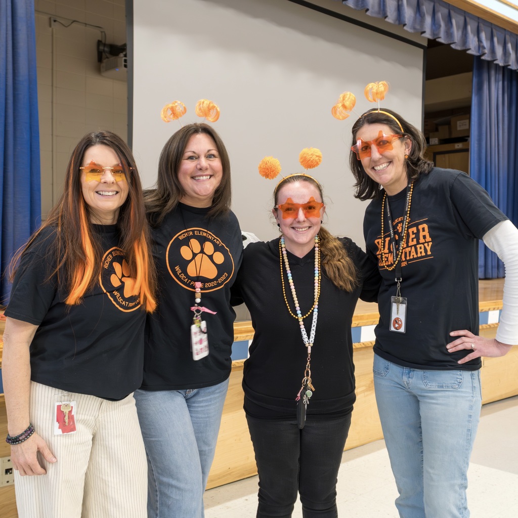 Four Richer School staff members in black t-shirts stand together in a classroom, wearing orange glasses and pom-pom headbands.