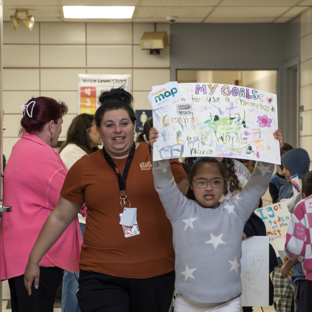 A group of Richer staff and students in a hallway; a student holds up a handmade sign with drawings and text. A Richer staff member walks next to her, smiling.
