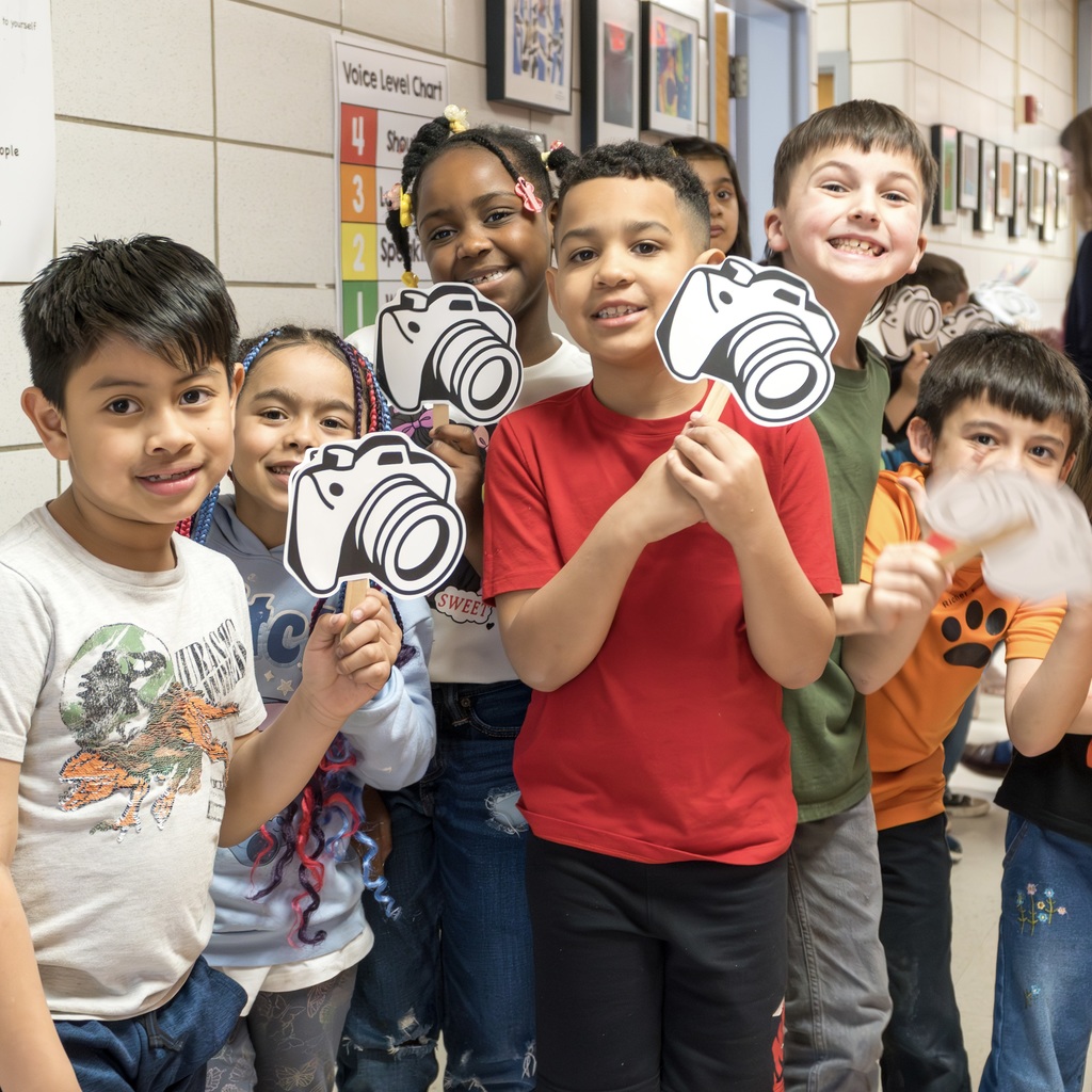 A group of Richer School students stands in a room, each holding a black-and-white camera cutout.