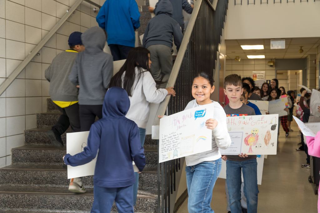 Children in a school hallway at the Richer School hold signs as they walk up a staircase, some with hoods up.