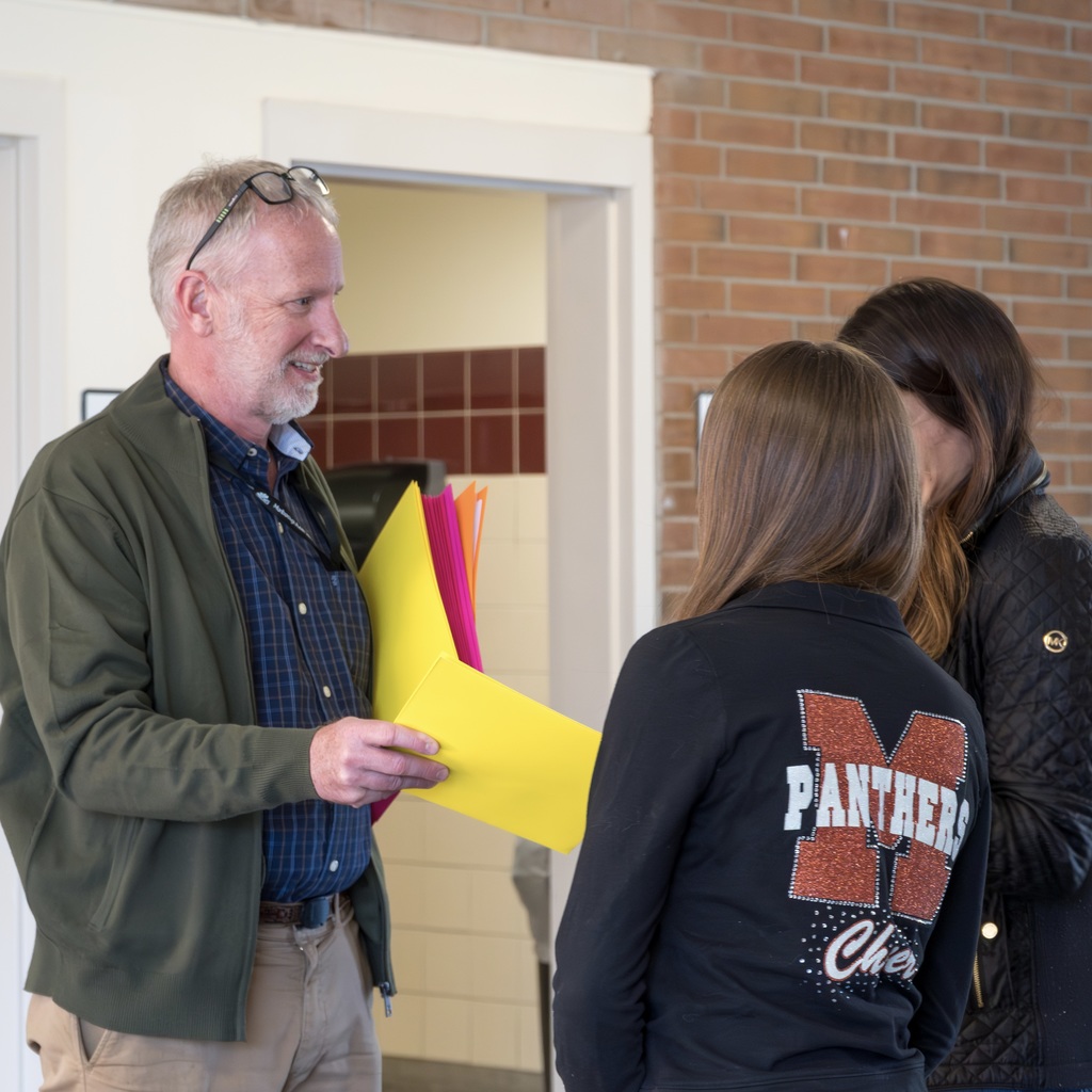 A man wearing glasses holds folders as he speaks to a parent and a student. The student wears a black Marlborough Panthers sweater.