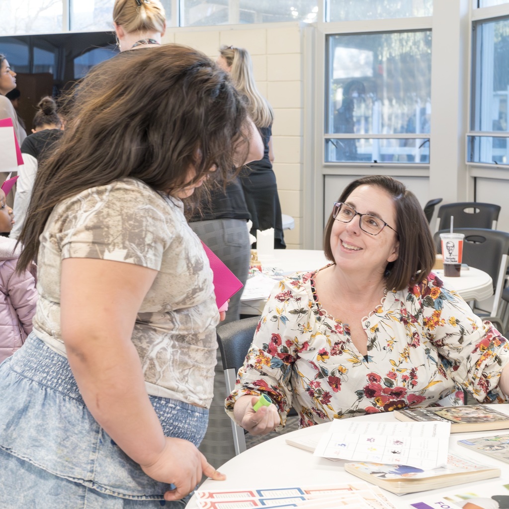 Two individuals talk in a room with windows; one is a teacher seated at a table with papers and books, while the student is standing.