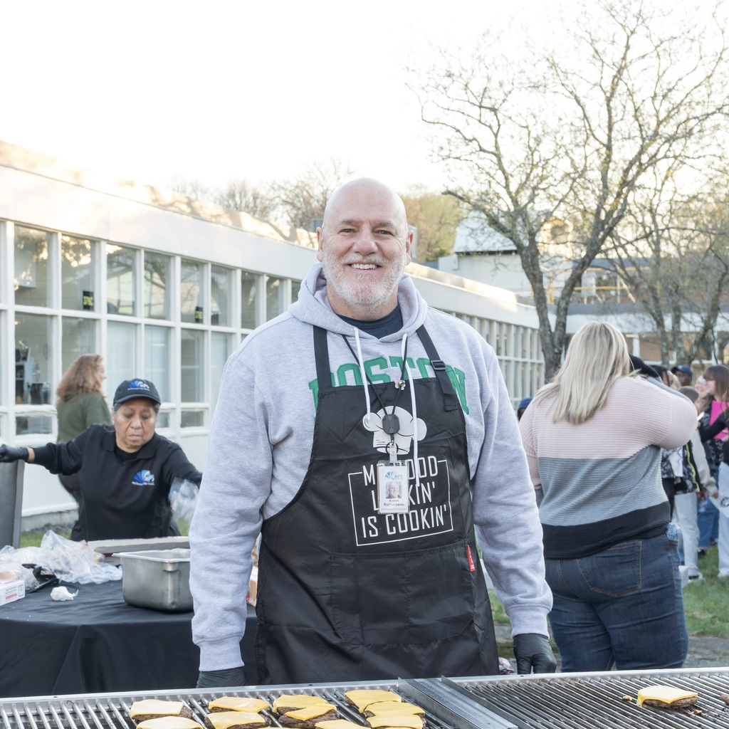 Whitcomb School Assistant Principal stands at a grill with food. Behind, two people prepare food at a table. The Whitcomb School building with glass walls in the background.