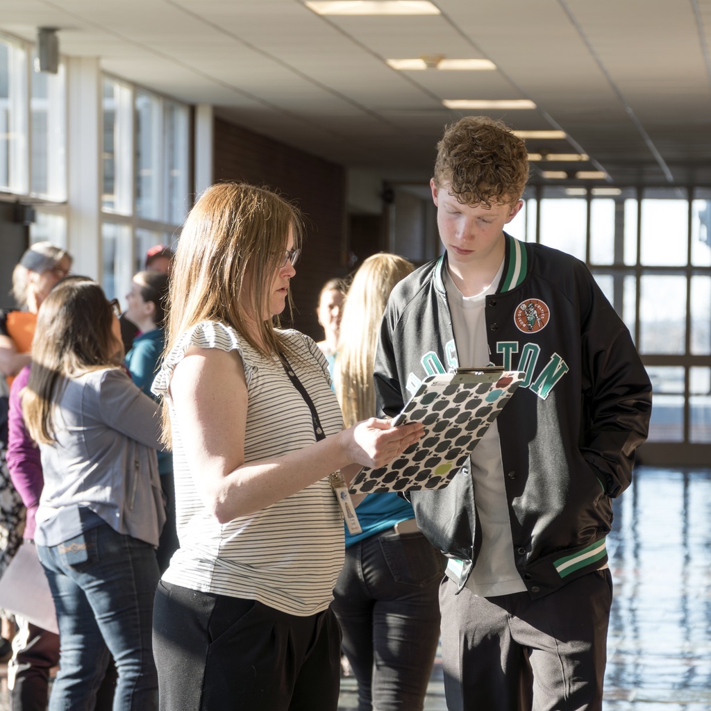 A woman and a man looking at a document, standing in a hallway with other people.