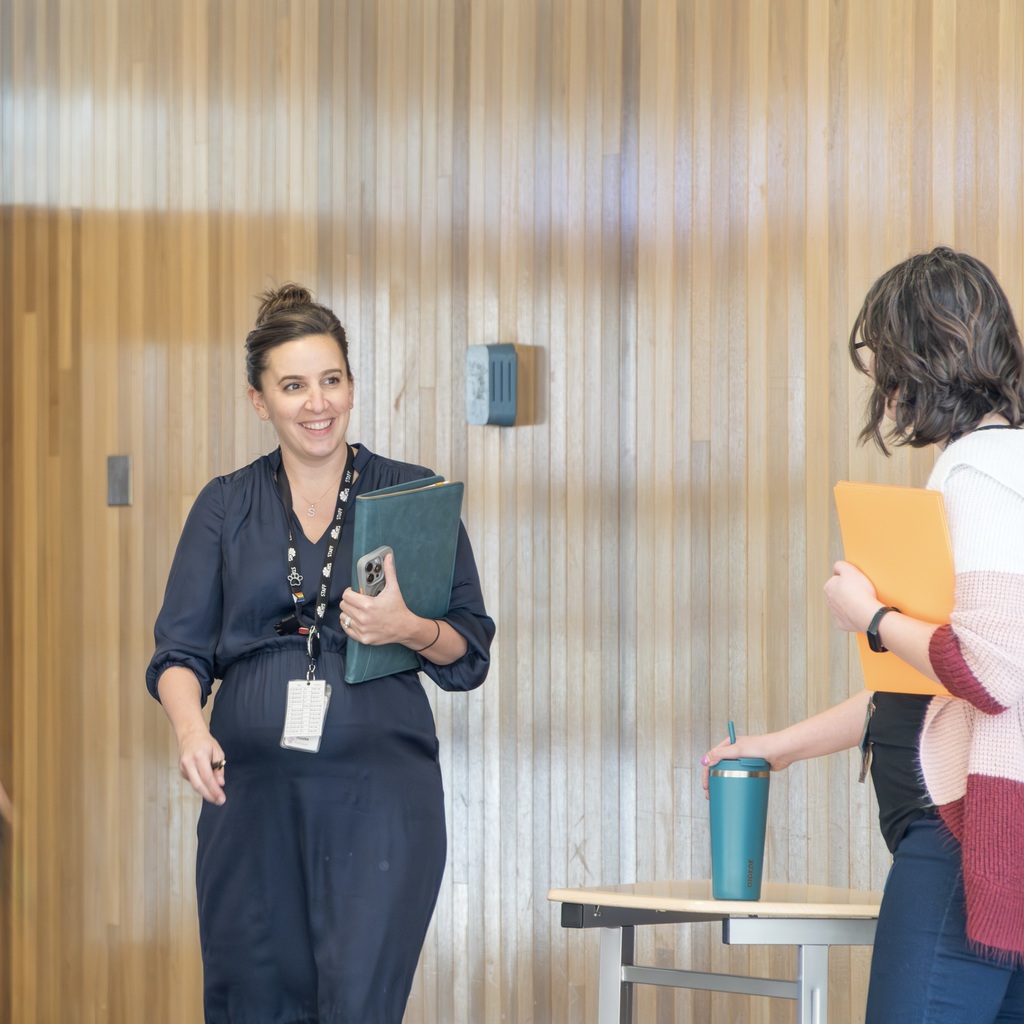 Whitcomb School Principal and another staff member stand in a room with wooden walls. Principal House holds a folder and smiles; the other holds a folder and a cup.