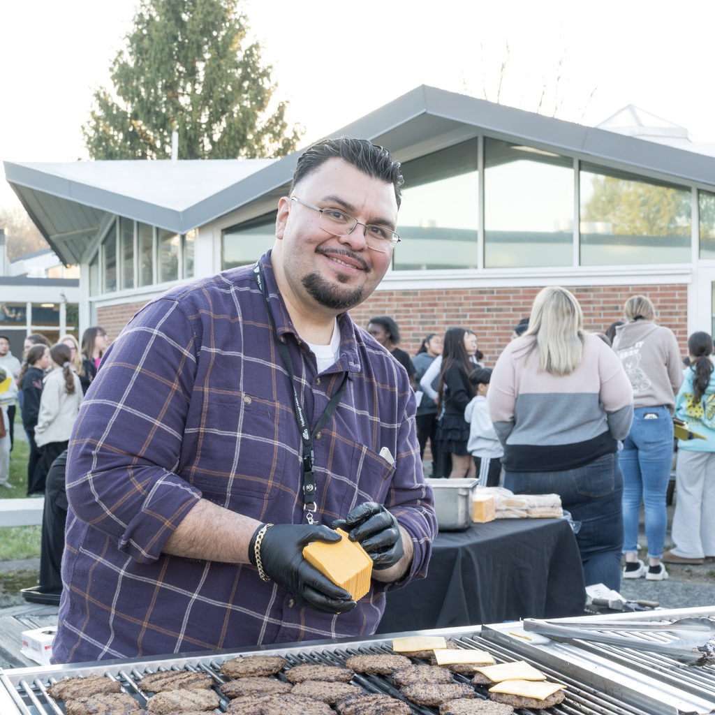 An MPS staff member with glasses and a plaid shirt grills burgers outside a building. He holds a block of cheese slices.