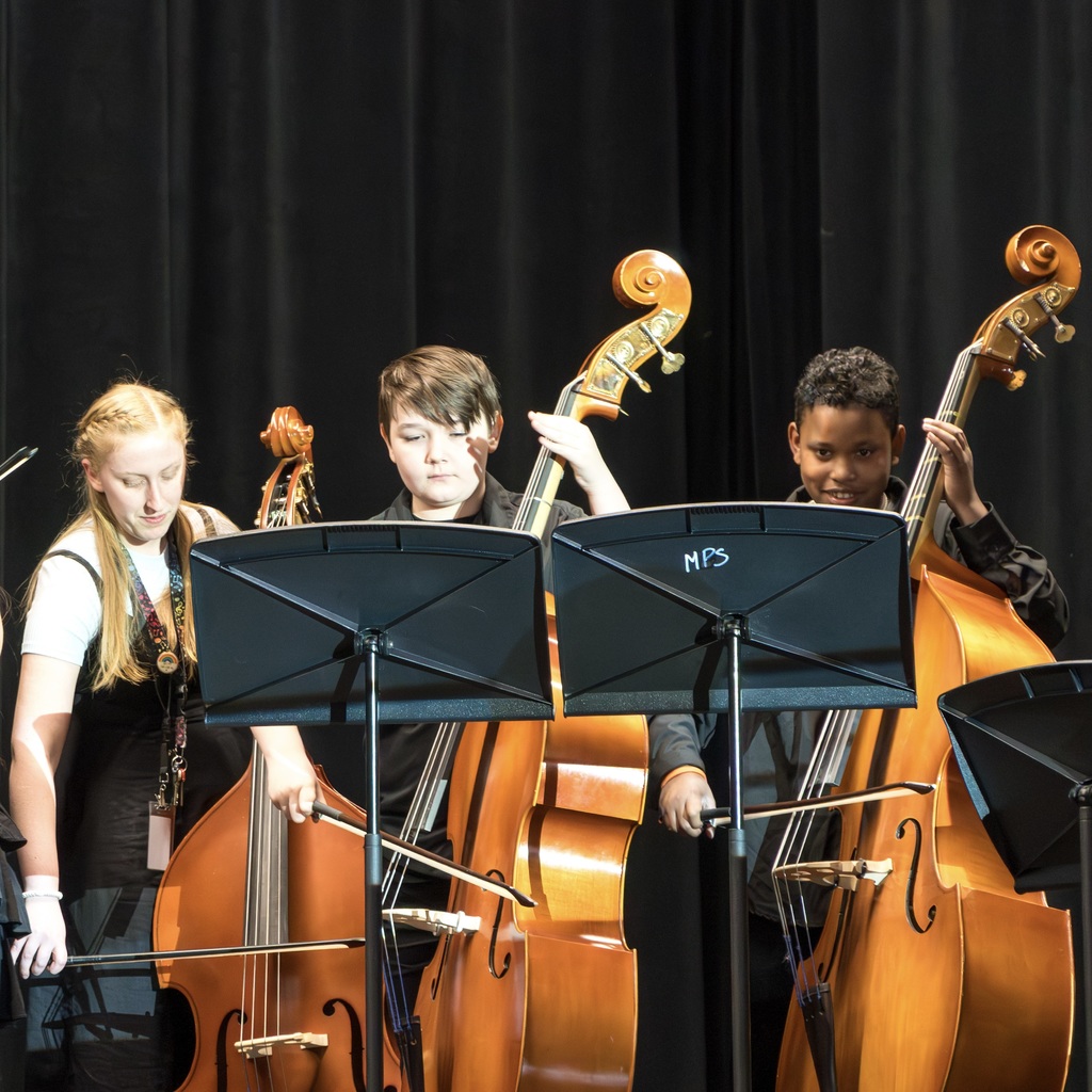 Two young Whitcomb School musicians and their teacher perform on cellos on a stage. They stand in front of music stands with black covers.
