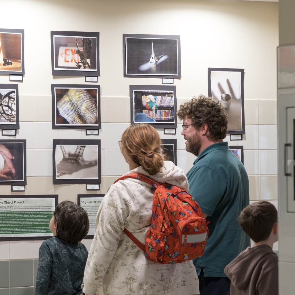 People in a corridor with framed images on a wall, a woman in white with a backpack, a man in green.