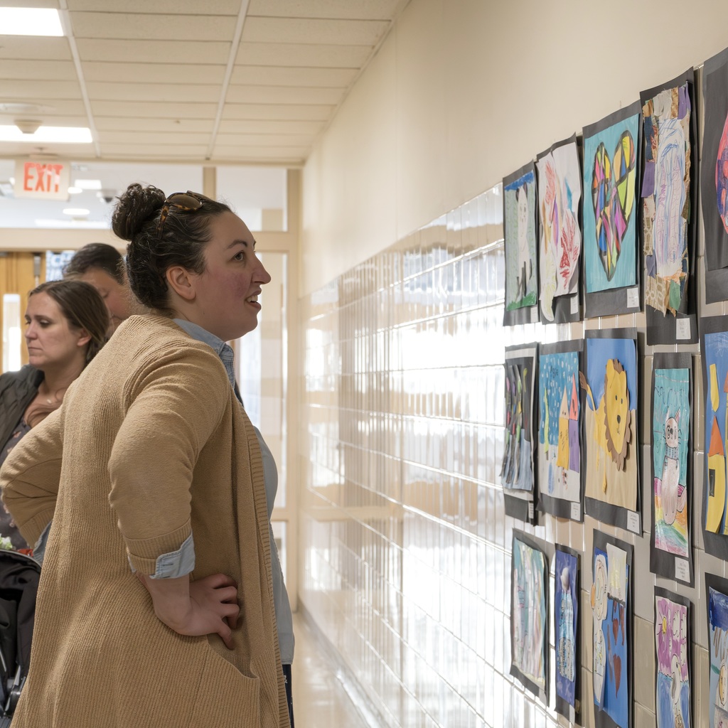Three individuals in light-colored coats walk past wall-mounted artwork in a hallway with fluorescent ceiling lights.