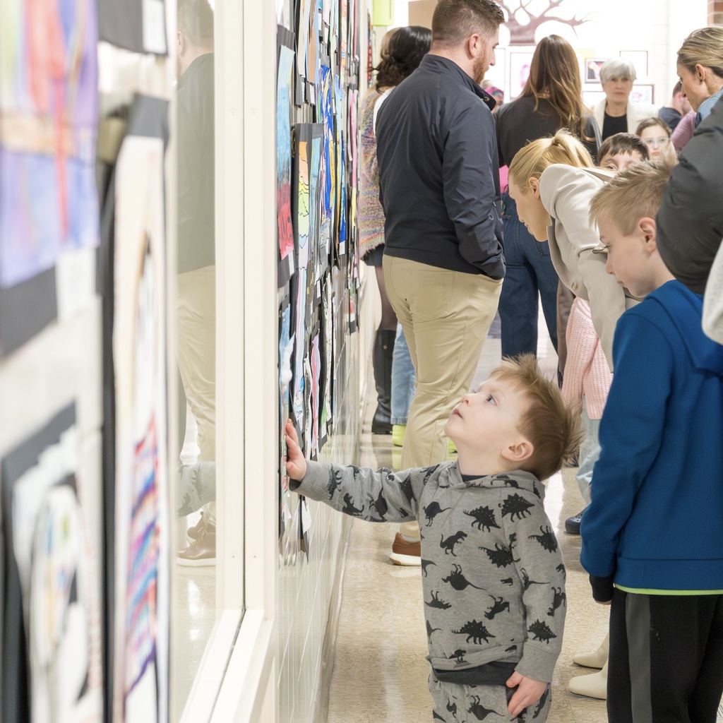 A group of people, including children, are gathered indoors around a wall adorned with colorful artwork.