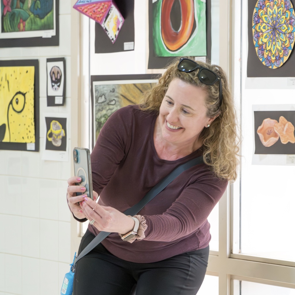 Woman with sunglasses on head taking photo of artwork in a room with framed pictures on the wall.