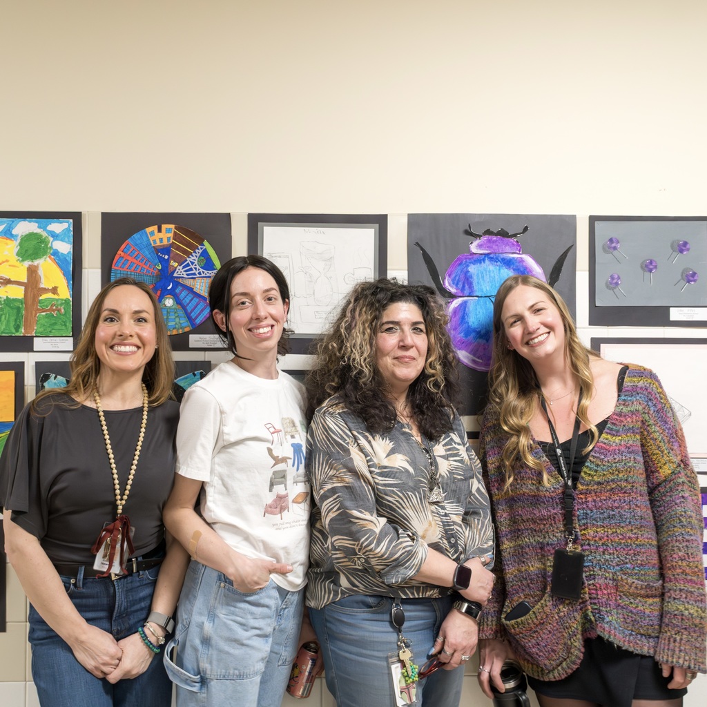 Four women stand together, smiling for a photo in front of a wall with multiple framed artwork.