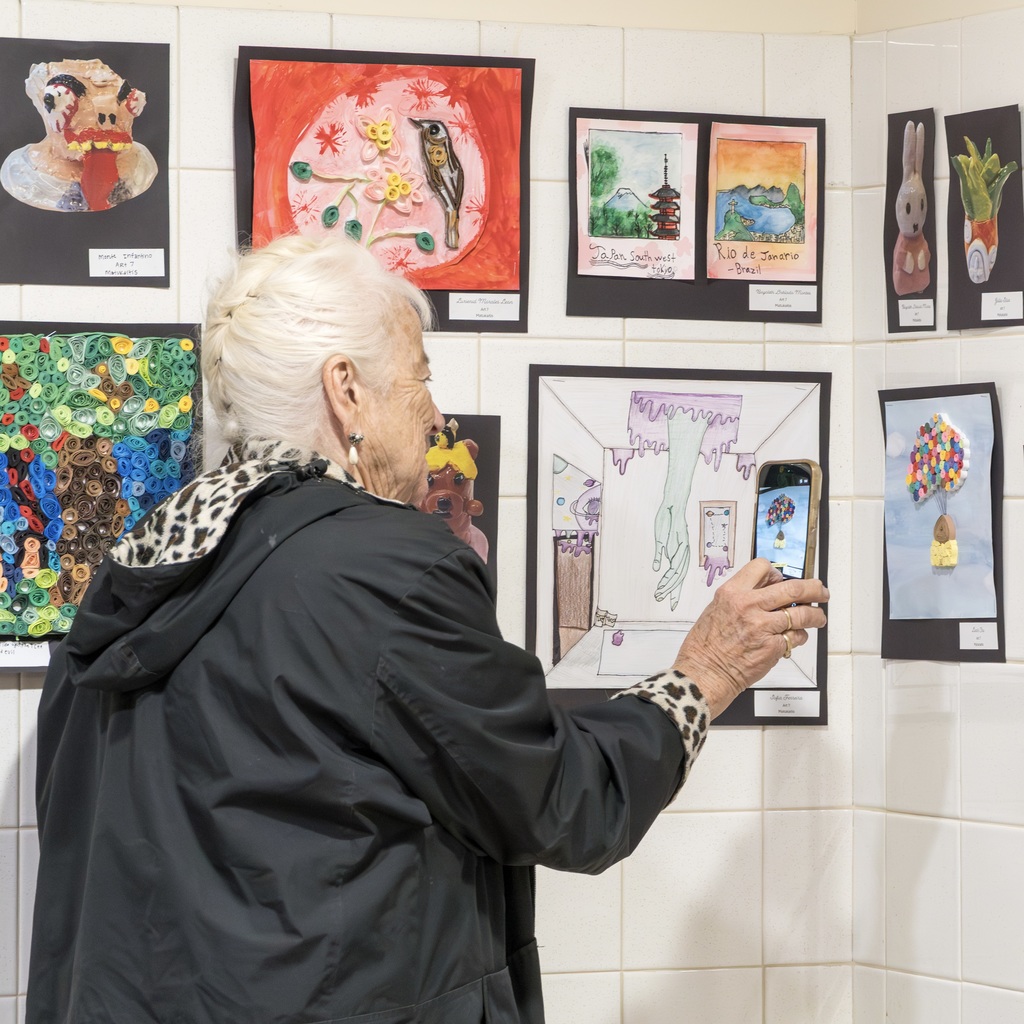 An elderly person takes a photo of art displayed on a wall. The art includes drawings of various subjects.