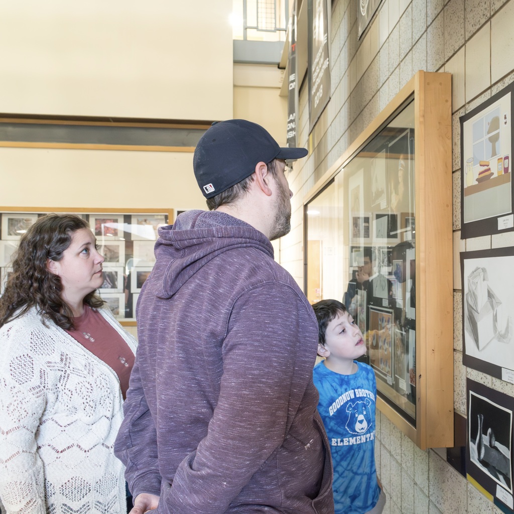 A man, woman, and child view artwork on a gallery wall with framed pictures. The man wears a hat, the woman a white sweater, and the child a blue shirt.