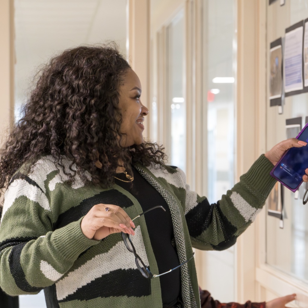 A person with curly hair wears a green-and-white sweater, holding glasses. They gesture to the right, facing a glass wall.