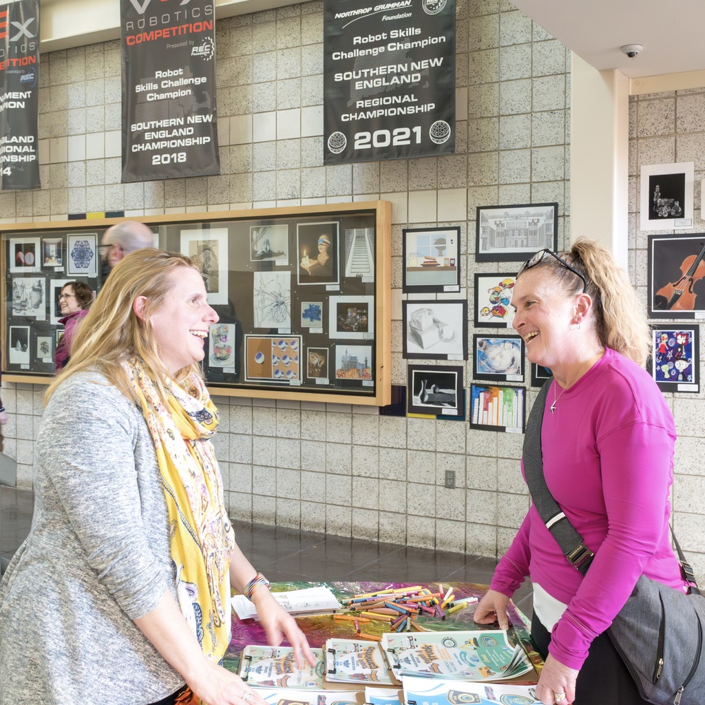 Two women stand at a table filled with items. Behind them, a display board shows various pictures and banners.
