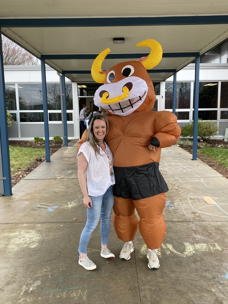 A woman in a white shirt, blue jeans, and white shoes stands next to a mascot in an orange costume with horns.