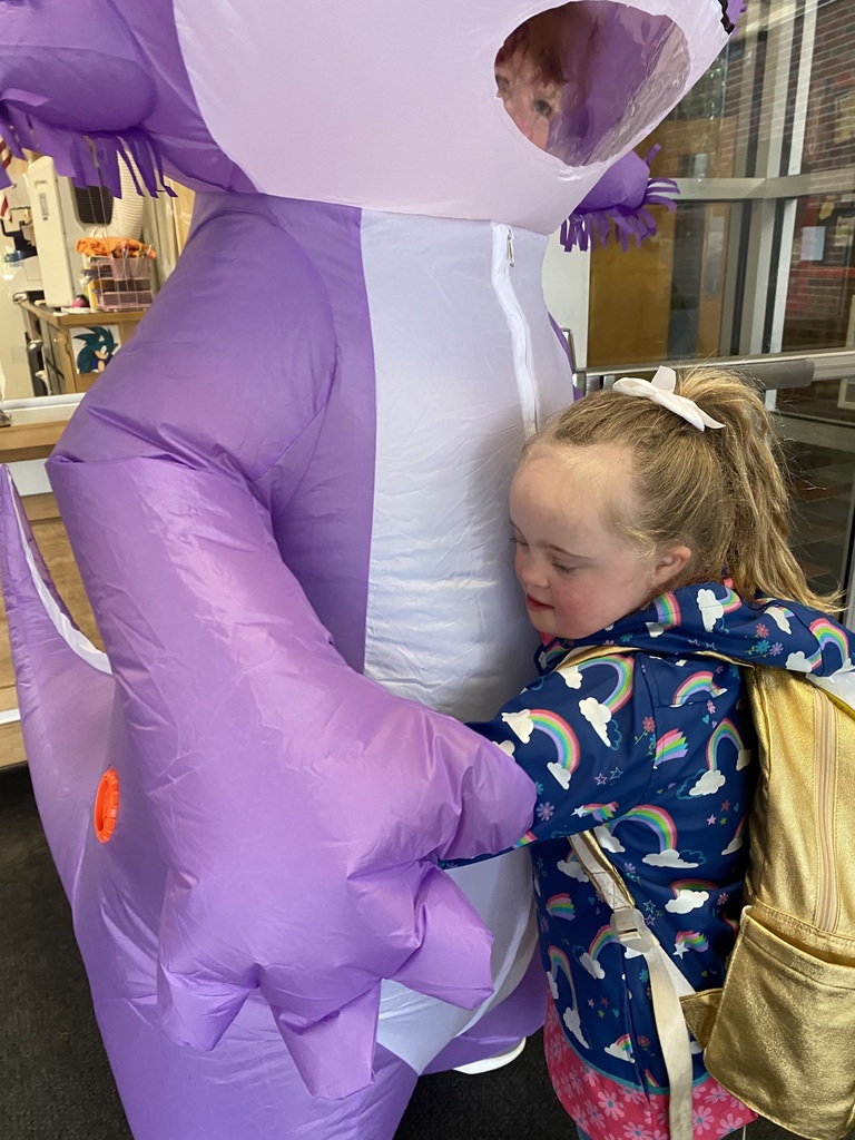 A young girl in a blue jacket stands close to a large purple balloon animal with a visible person inside.