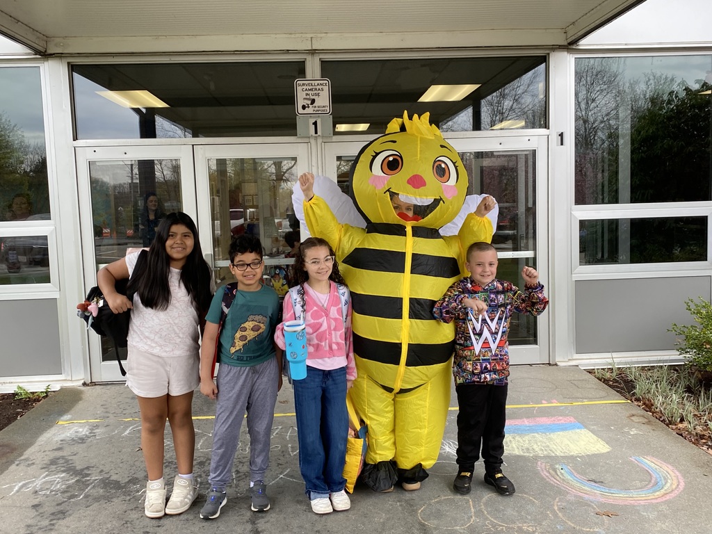 Four children and a bee mascot stand in front of a building with glass doors.
