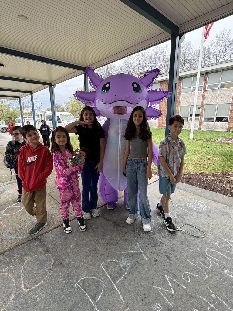 Six children, some in costumes, stand under a school shelter with a purple mascot. A building is in the background.