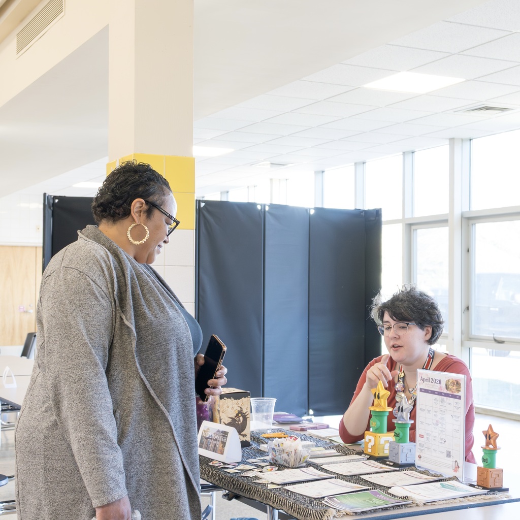 Two individuals at a table with papers and items; one, standing and holding a phone, talks to the other, sitting at the table. Bright, well-lit room with windows and a wooden wall in the background.