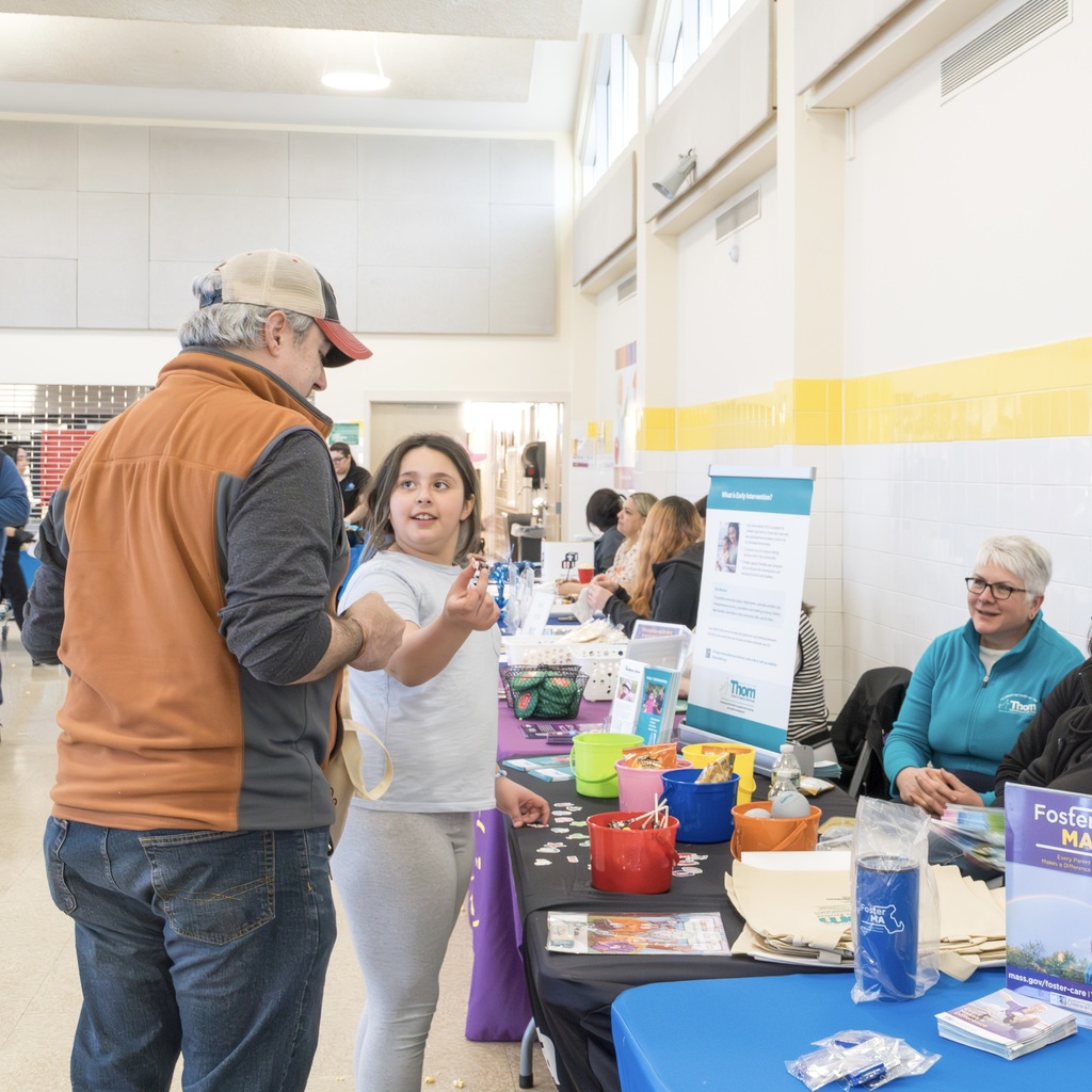 A man and a girl interact at a community event. A table with various items is behind them.