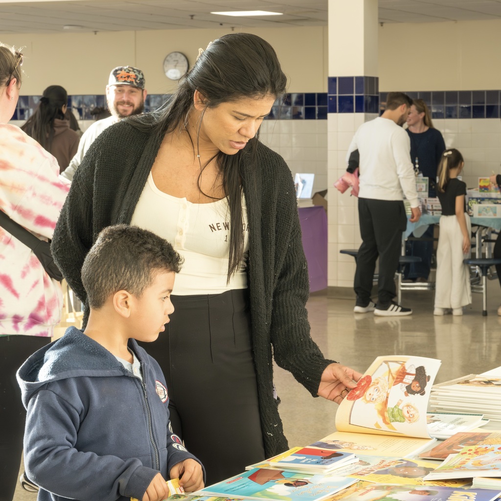 A woman with a child at a book fair. People in a tiled room. Books on a table.