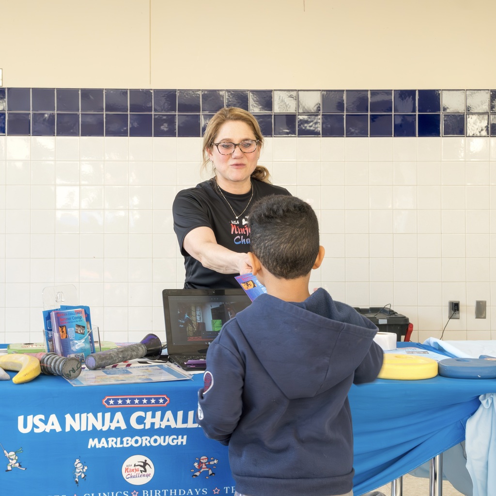 A woman standing behind a table. A boy in a blue jacket stands across from her.