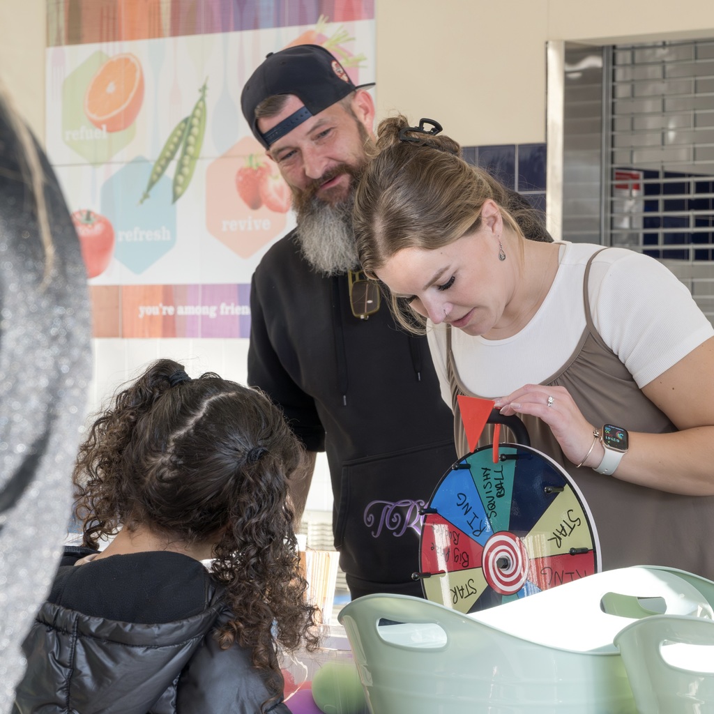 Three people at an event: a man, a woman, and a child. The woman stands looking down at a prize wheel.