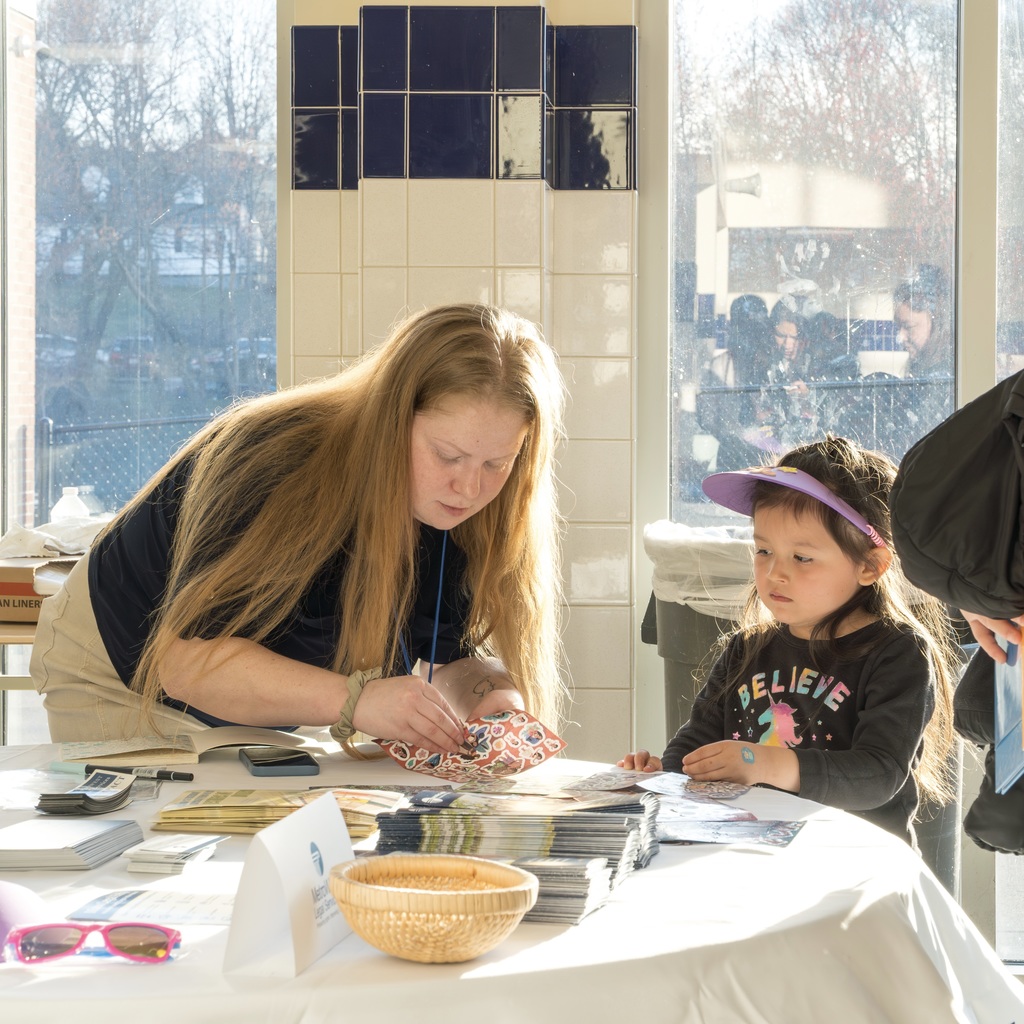 A woman with long hair is at a table, helping a child with a sticker sheet. The table has a basket, sunglasses, and books.