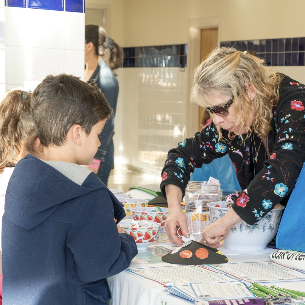 A woman helps a child at a table with bowls and papers. Behind them, a person stands by a door.