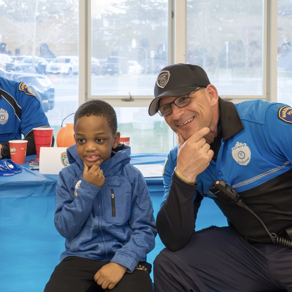 A police officer and a child sitting at a table smile and pose. Outside, cars are parked.