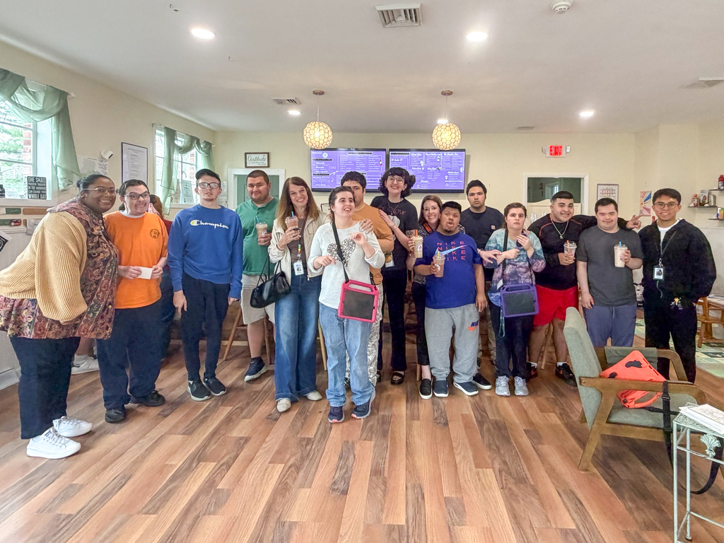 A group of MHS students and staff stand together indoors, wearing casual clothes. Some hold cups. Behind them, a monitor displays the restaurant menu.
