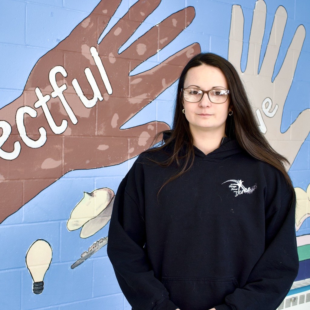 Substitute teacher Antoinette Brown, wearing glasses, stands in front of a blue mural with hand prints and the word "Respectful."