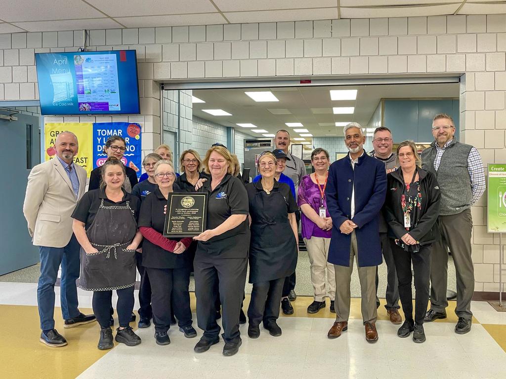 A group of MPS staff and faculty, in uniform or business casual attire, stand together, smiling. Some hold a plaque. A screen and lights are on the wall behind them.