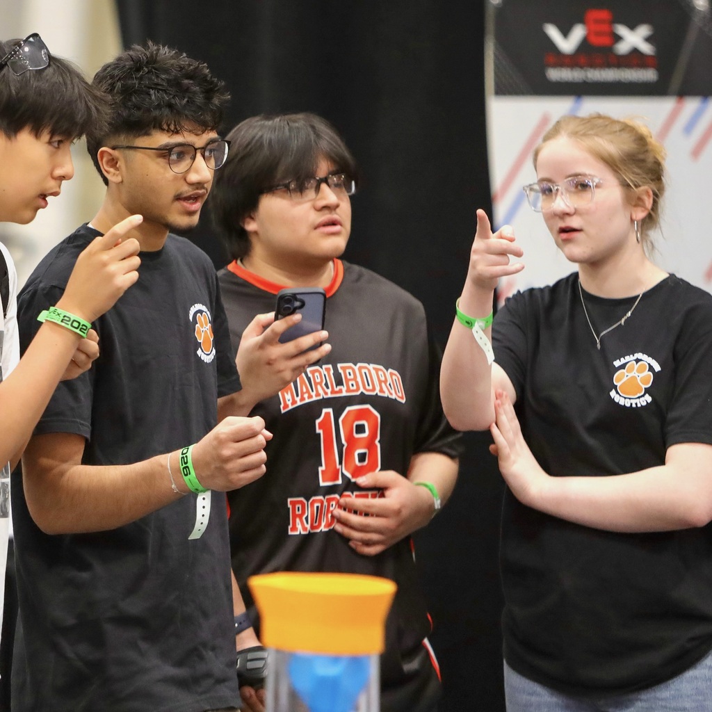Three MHS Robotics team members and another young individual, one holding a phone, stand and talk.
