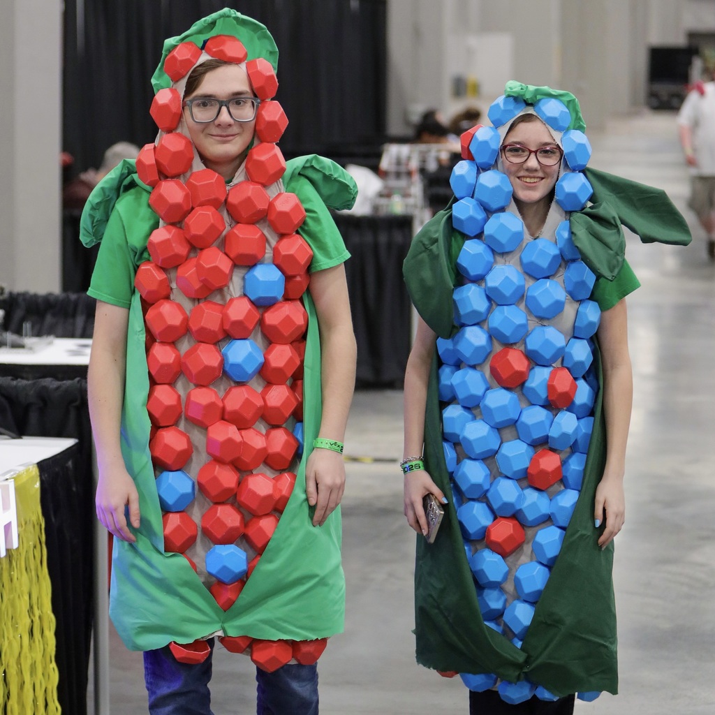 Two people wear costumes covered in blue and red shapes in an indoor area with tables.