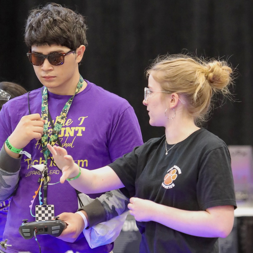 Two individuals, one in a purple shirt and the other in black, are facing each other and talking with their hands extended. Background blurred.