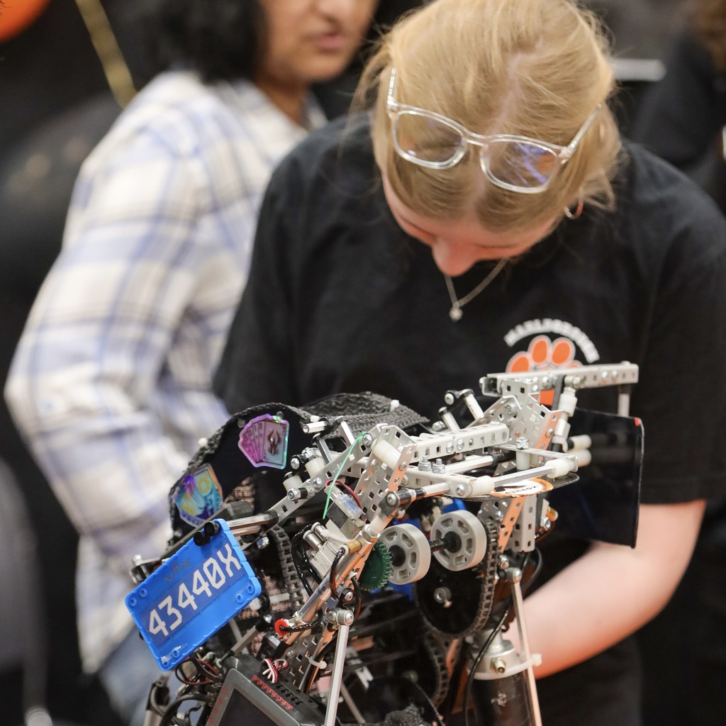 MHS female student wearing glasses and a black shirt inspects a mechanical device labeled 4344OX. Behind her, a person in a white-and-blue plaid shirt stands.