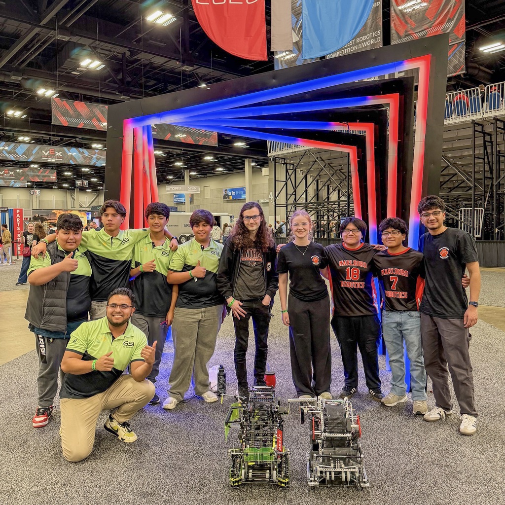 Nine individuals are lined up, posing for a photo. Some are kneeling, and others are standing behind a large, colorful frame.