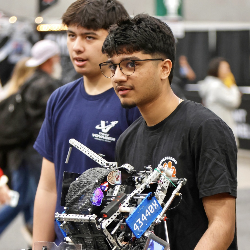 An MHS robotics team member and another individual, one in glasses, hold a large mechanical robot outdoors. Several people walk in the blurry background.