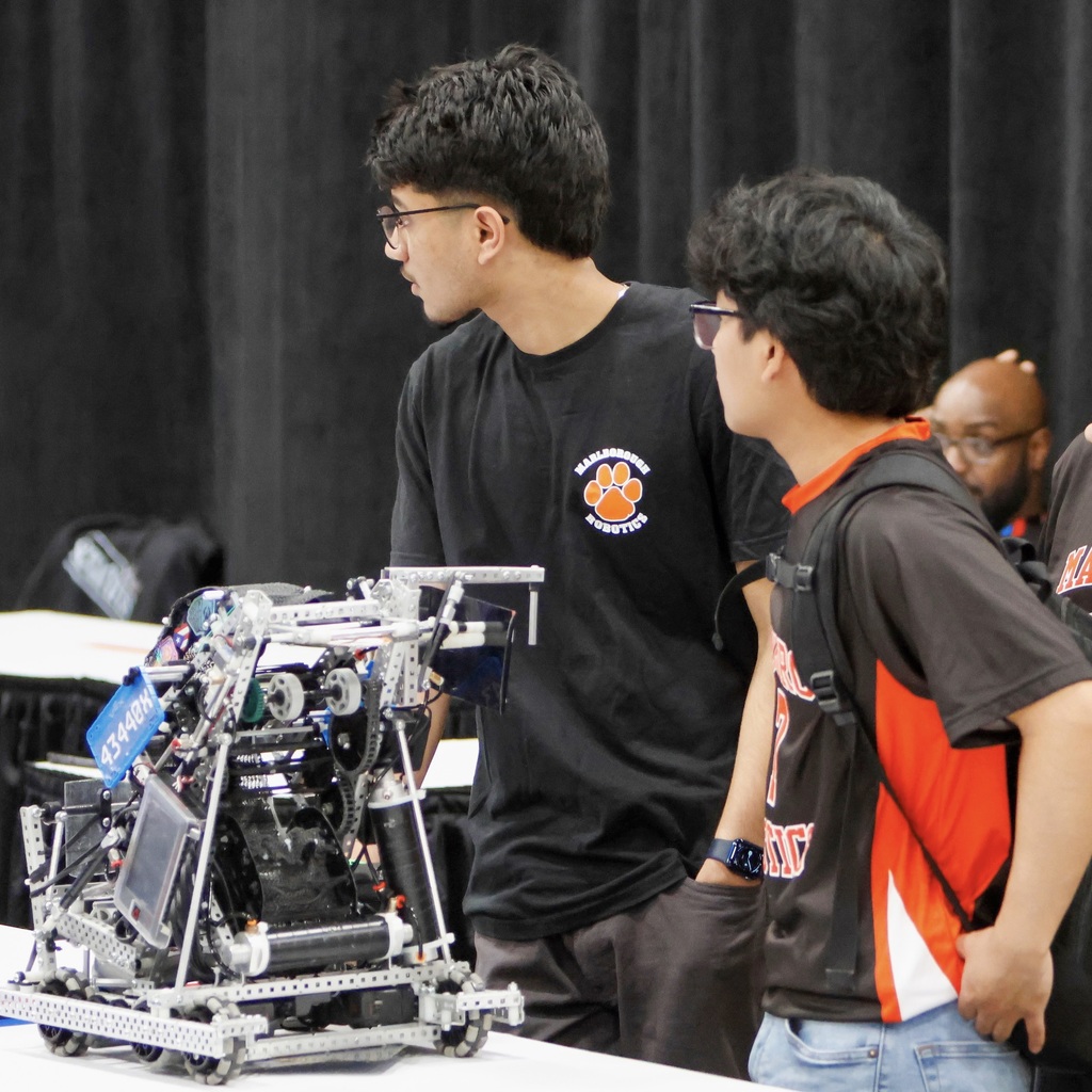 Two MHS Robotics team members are standing around a robotic vehicle on display at an indoor event.