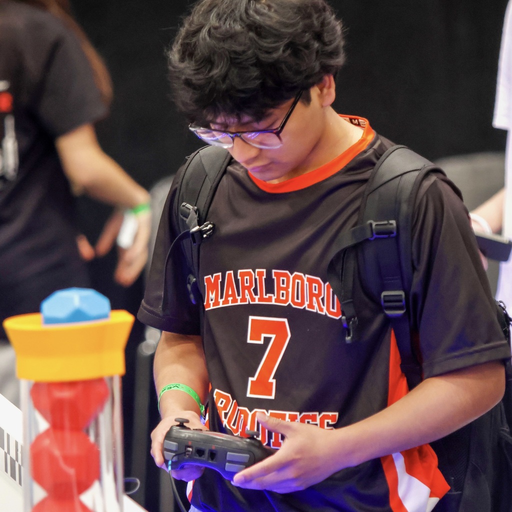 An MHS Robotics team member wearing a black shirt with "MARLBORO" and number 7 stands with a backpack, holding a game controller.