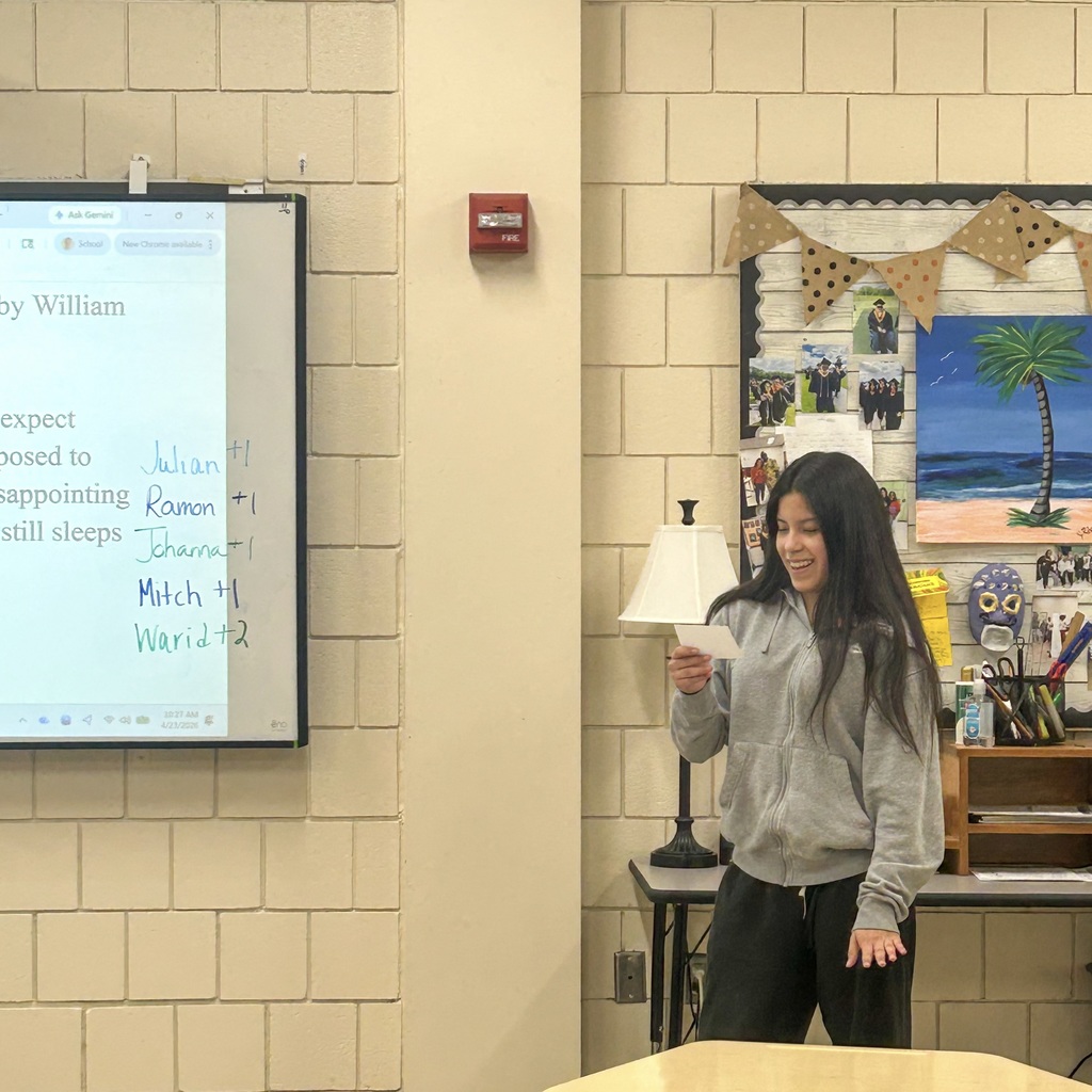 An MHS female student holding paper, standing at a desk, with a lamp, framed artwork, and a classroom screen showing text.