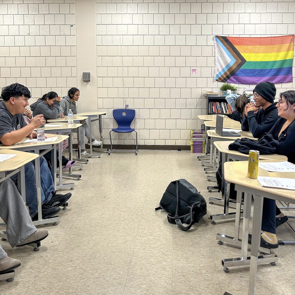 Group of MHS students in a classroom. Desks and chairs are arranged in rows facing each other. A rainbow flag hangs on a white brick wall.