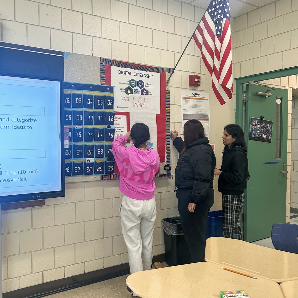 Three people in a classroom stand facing a wall; two are writing on a board. A screen displaying information to their right, an American flag above them.