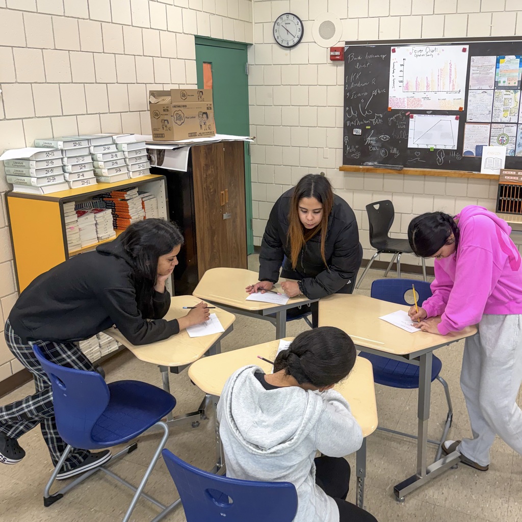 Four MHS students in a classroom with a blackboard. One sit at desks, writing. Three stand and write.