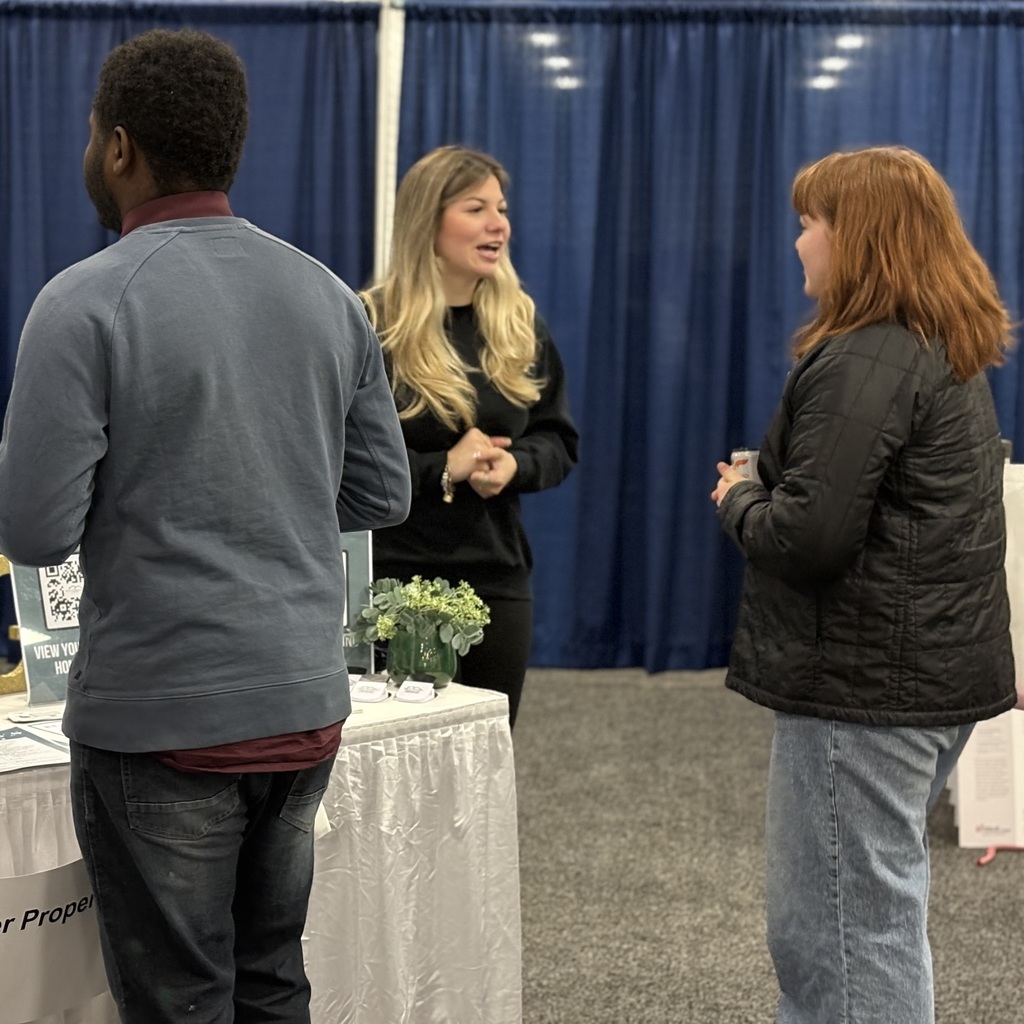 An MHS alum speaks with a woman near a table; a man stands nearby, facing away from the camera.
