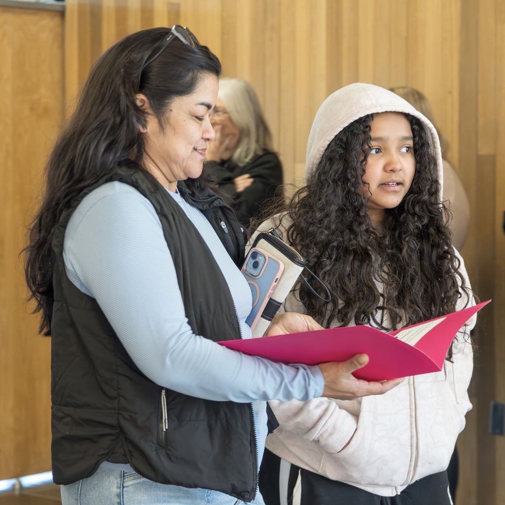 An MPS female student and their mom stand in a hallways, one holding a folder open. The mom on the left wears a vest and holds a folder.