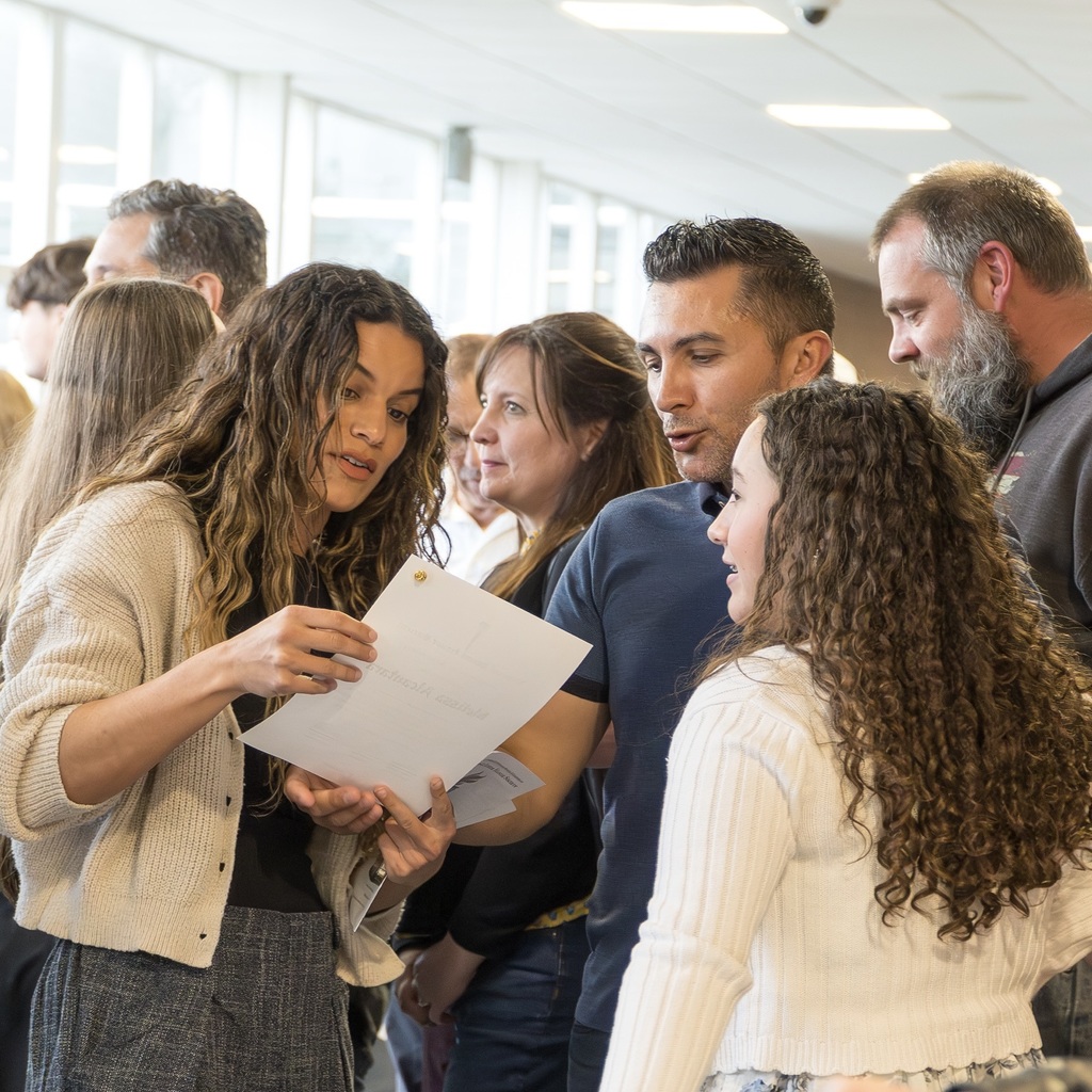 A group of people standing in a hallway, a woman holding a paper looks at it while a student and family member look on.